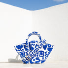 Marabu basket bag in blue Symbols print — natural cotton with shoulder strap, lifestyle shot against white Greek wall and blue sky