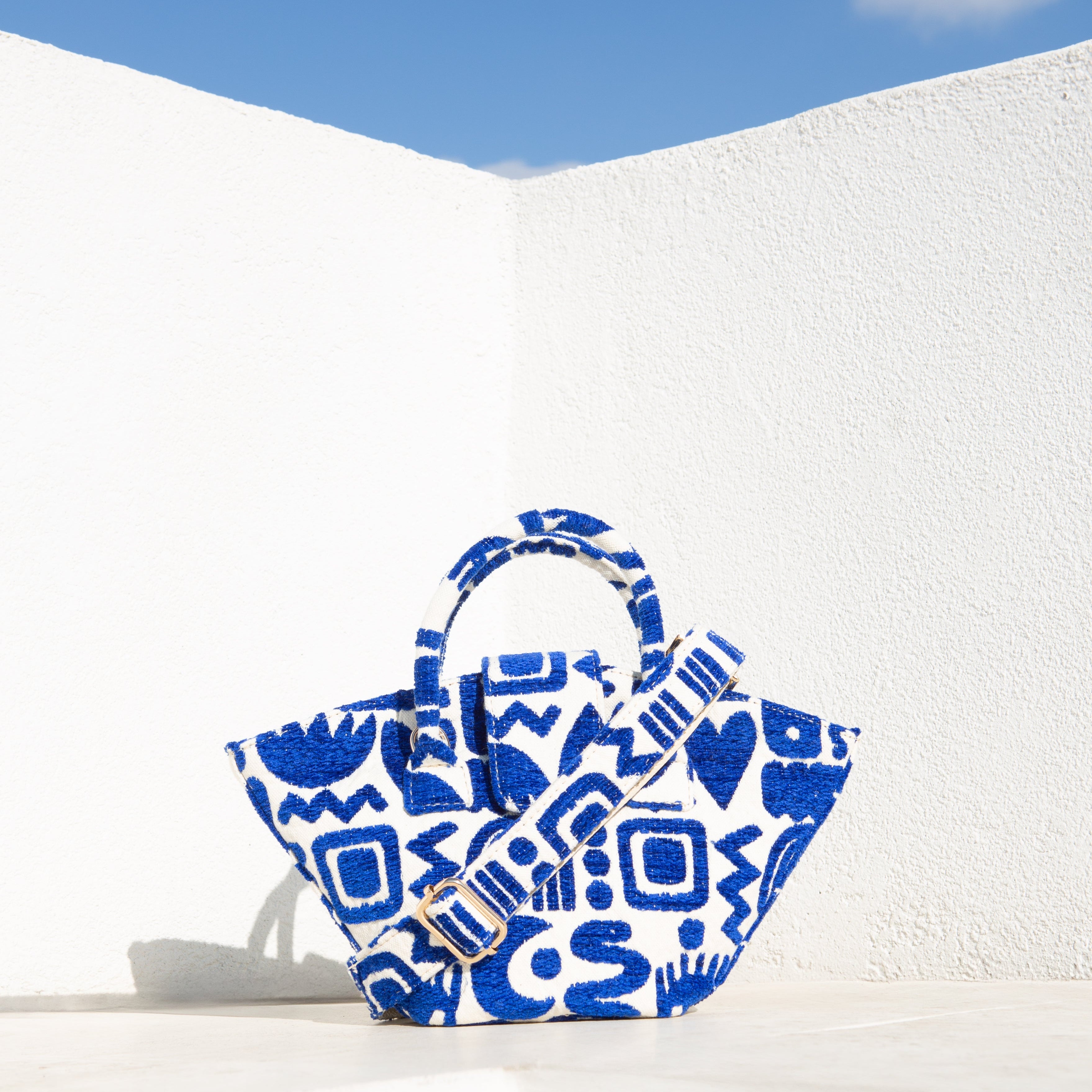 Marabu basket bag in blue Symbols print — natural cotton with shoulder strap, lifestyle shot against white Greek wall and blue sky