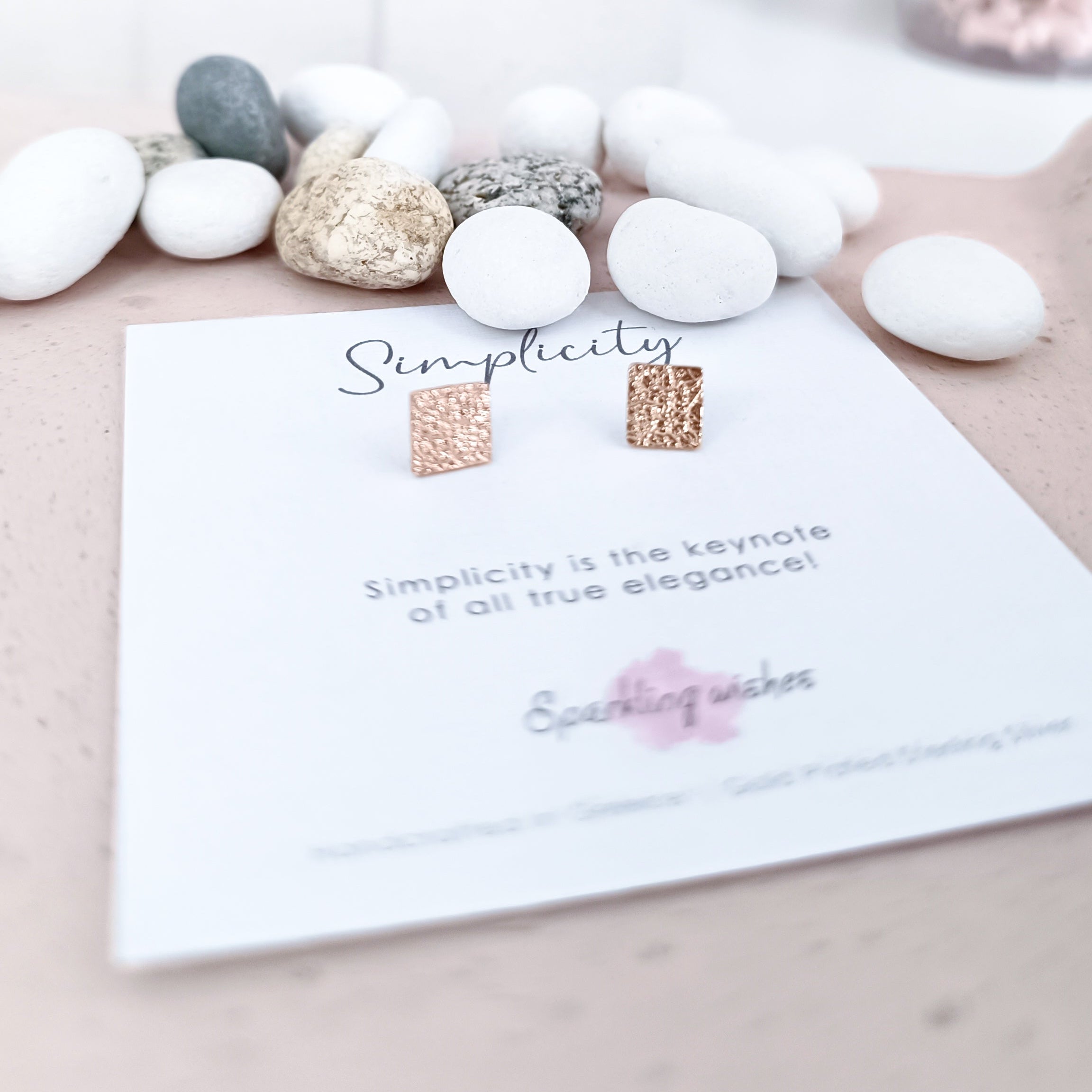 Pair of earrings on a card with 'Simplicity' text, surrounded by decorative stones.